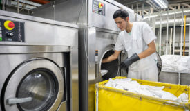 Focused man working at an industrial laundry service loading washing machine wearing protective gloves - Business industry concepts